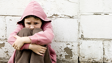 Sad child sitting outdoors against a wall.